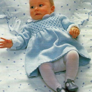 May include: A baby wearing a light blue knitted dress with a white collar and white tights. The baby is lying on a white blanket with a blue and white pattern.
