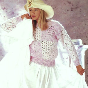 May include: A pale pink and white knitted jumper with openwork patterns. The jumper features long sleeves and a ribbed hem. The model is wearing a wide-brimmed hat with a flower and a white skirt. The jumper is a soft, feminine design.