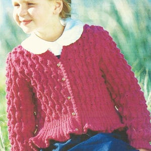 以下が含まれることがあります： 白い襟とボタンが付いたピンクのニットカーディガンを着た若い人。カーディガンはテクスチャのあるデザインで、濃い青色のスカートの上に着ています。