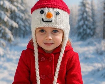 May include: A child wearing a red coat and a knitted snowman hat with a red pom-pom, black button eyes, and an orange nose. The hat has white ear flaps with white pom-poms at the ends. The background is a snowy winter scene.