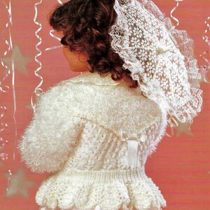 May include: A white knitted cardigan with a ruffled hem and a bow at the back. The cardigan is worn over a white dress and the girl is wearing a white lace hat.