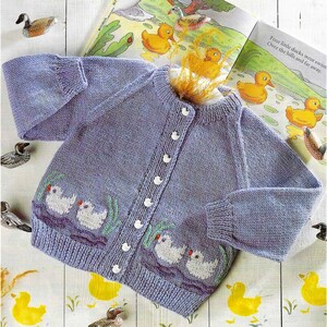 May include: A light purple knitted baby cardigan with white buttons and embroidered white ducks on the front. The cardigan has a ribbed collar and cuffs. The text "Four little ducks went swim Over the hills and far away." is visible in the background.