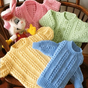 Puede incluir: Una colección de coloridos suéteres y un gorro de bebé tejidos a mano en rosa, amarillo, verde y azul. Los suéteres presentan diseños de punto trenzado y texturizado. También hay un peluche de conejo.