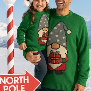 May include: A father and child in matching green Christmas jumpers with a gnome design. The jumpers feature a grey hat with white polka dots, a brown beard, and a red gift. The child wears a Santa hat. A sign reads "NORTH POLE".