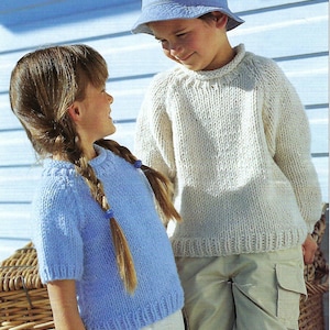 May include: Two children in hand-knitted sweaters and shorts. The girl wears a light blue short-sleeved sweater, and the boy an off-white sweater and a blue bucket hat. They are near a wicker basket. The sweaters appear to be handmade.