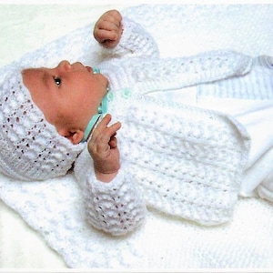 May include: A white crocheted baby hat and cardigan set. The hat has a lacy design and the cardigan has a button closure.