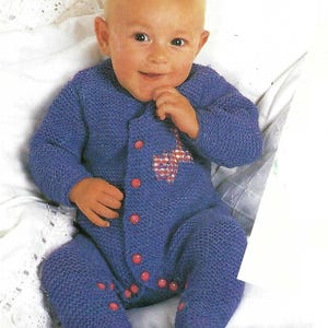 May include: A knitted baby onesie in a purple shade with pink buttons and a small red and white patterned appliqué on the front.