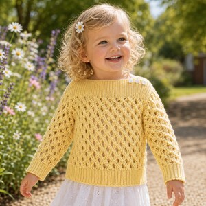 May include: A yellow knitted sweater with a diamond pattern and three white buttons on the shoulder. The sweater has long sleeves and a ribbed hem. The child is wearing a white skirt and a flower in their hair.