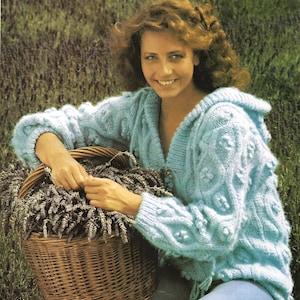 Könnte beinhalten: Eine Frau trägt einen hellblauen Strickcardigan mit Kapuze und hält einen Weidenkorb voller Lavendel. Der Cardigan hat ein strukturiertes, noppenartiges Design.