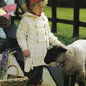 May include: A child wearing a cream-colored, hooded cable-knit coat with wooden toggle closures and matching mittens. The child is also wearing a red polka-dot headscarf, red boots, and plaid pants. A sheep stands nearby.