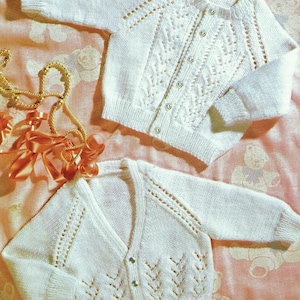 May include: Two white knitted baby cardigans. One has a round neck and the other a V-neck. Both have long sleeves and button fastenings. The cardigans feature decorative knit patterns and are displayed on a pink patterned surface.