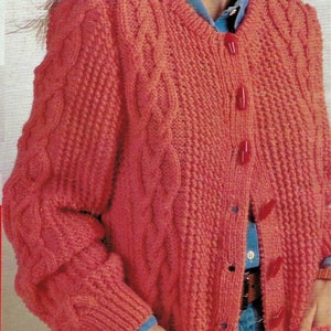 May include: A pink knitted cardigan with a cable knit pattern and button closure. The cardigan has pockets on the front and is worn over a blue denim shirt.