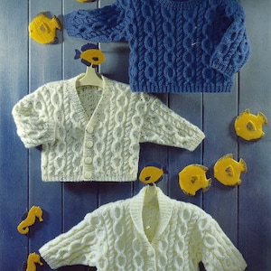 May include: Three knitted baby sweaters, one blue, two white, with a cable knit pattern. The sweaters are hanging on hangers against a blue wooden background.
