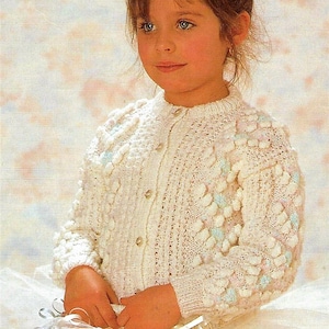 May include: A white long-sleeved knit cardigan featuring a textured bobble pattern with subtle pastel blue and pink accents. The garment has a round neckline, ribbed cuffs, and a front button closure with small, light-coloured circular buttons.