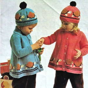May include: Two children wearing knitted cardigans and matching hats with a turtle design. The cardigan on the left is blue and the cardigan on the right is pink. The hats have a brown, white, and blue turtle design.