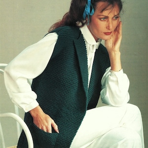 Peut inclure: Un gilet tricoté vert foncé avec une poche, porté sur une chemise blanche à col et un pantalon blanc. Le gilet a un design long et ouvert sur le devant. Le modèle est assis sur une chaise blanche.