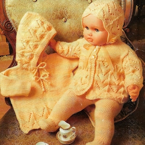 May include: A yellow knitted baby doll outfit with a matching bonnet. The outfit features a cardigan with a button closure and a lace pattern. The doll is sitting on a chair with a floral pattern.