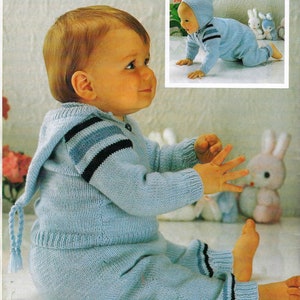 Può includere: Un bambino che indossa un maglione di lana blu chiaro con cappuccio e pantaloni abbinati. Il maglione ha strisce nere e blu sulle maniche e una lunga cravatta intrecciata al collo. Il bambino è seduto su una superficie bianca con un peluche nelle vicinanze.