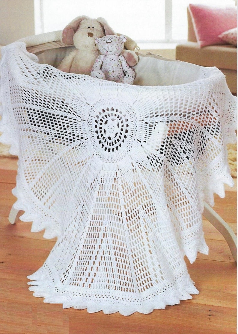 May include: White crocheted baby blanket with a delicate lace pattern and scalloped edges. The blanket is draped over a white wicker bassinet with two stuffed toys inside.