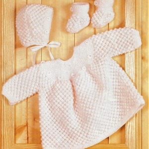 May include: A white knitted baby outfit set, including a dress, a bonnet, and booties. The dress has long sleeves and a round neckline. The bonnet has a tie at the back. The booties are small and have a pointed toe.