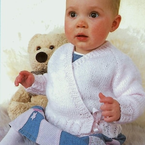 May include: A baby wearing a light pink knitted wrap sweater with a matching bow. The baby is also wearing blue and pink patchwork pants and white tights. A beige teddy bear is in the background.