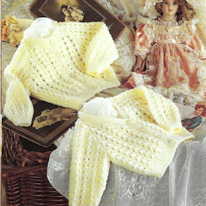 May include: Two pale yellow knitted baby cardigans with a delicate openwork design are displayed on a lace-covered surface. The cardigans have long sleeves and small button closures. A vintage doll and framed photograph are in the background.