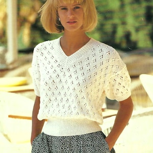 Puede incluir: Un suéter blanco de manga corta con cuello en V y un patrón de punto calado. El suéter tiene una cintura acanalada y se combina con una falda estampada en blanco y negro. El estilo general es casual y vintage.