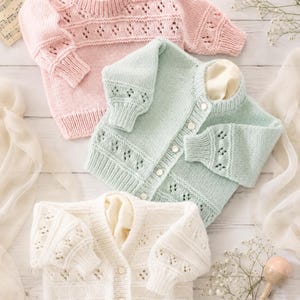 Puede incluir: Tres suéteres de bebé de punto en colores pastel. Un jersey rosa claro, una rebeca azul claro con botones y una rebeca blanco roto con botones. Cada suéter tiene un delicado diseño calado en las mangas y el cuerpo. Los suéteres se exhiben sobre una superficie de madera clara.