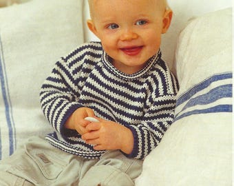 May include: A smiling baby wearing a navy and white striped sweater with a turtleneck collar and khaki pants. The baby is sitting on a white surface with a white pillow behind them.