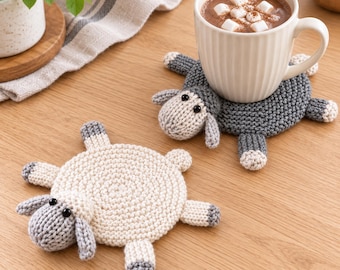 May include: Two round, hand-knitted coasters shaped like sheep. One coaster is cream-colored with a gray head and feet, while the other is dark gray with a light gray head and feet. Each features small black bead eyes and four short legs.