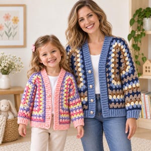 May include: Two crochet cardigans featuring a granny square pattern. The smaller cardigan is pink, yellow, and purple with white pearl-style buttons. The larger cardigan is blue, brown, and cream with white pearl-style buttons and a ribbed trim.