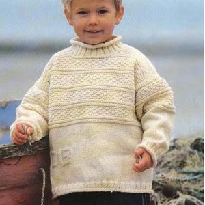 Peut inclure: Un jeune garçon portant un pull en tricot crème avec un motif texturé. Le pull a un col montant et des manches longues. Le garçon est debout devant un bateau en bois.