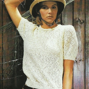 Puede incluir: Un top blanco de crochet con cuello redondo y mangas cortas. El top tiene un delicado patrón de calado. La modelo lleva un sombrero marrón con una correa para la barbilla.