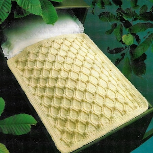May include: A crocheted baby blanket in a pale yellow colour with a diamond pattern. The blanket is lying on a cot.