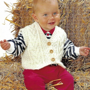 May include: A white knitted baby vest with a cable knit pattern and wooden buttons. The vest is worn by a baby sitting in a pile of hay.