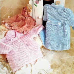 May include: Two knitted baby sweaters, one pink and one blue, with button closures. The sweaters are displayed on a white surface with a doll in the background.