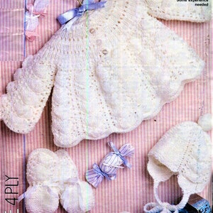 May include: White knitted baby cardigan, hat, and booties. The cardigan has a button closure and a lacy pattern. The hat has a pointed top and a tie. The booties have a bow detail. 46-48 cm (18-19 in). Some experience needed.