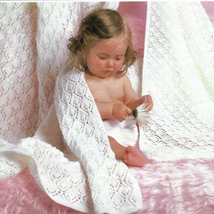 May include: A baby wrapped in a white, open-weave blanket with a diamond pattern. The baby is holding a white daisy and sitting on a pink, fluffy surface. The blanket is draped around the baby, creating a cosy and soft appearance.