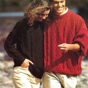 May include: A couple wearing matching cable-knit sweaters. One sweater is black, the other is red. Both sweaters have a textured pattern. The couple is also wearing khaki pants. The background is blurred.