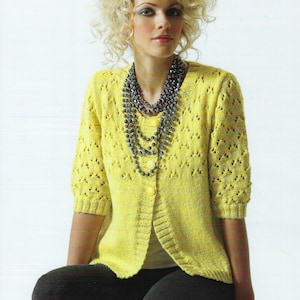 May include: A yellow knitted cardigan with a delicate openwork pattern and a button fastening. The cardigan has three-quarter sleeves and is worn over a white t-shirt. The model is wearing a layered necklace with dark beads.