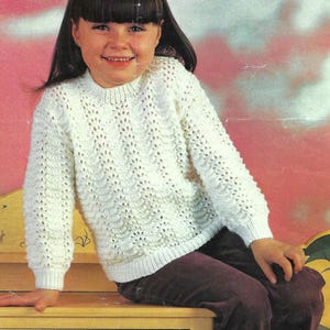 May include: A cream-colored, knitted sweater with a textured pattern. The sweater has a crew neck and long sleeves. The person in the image is wearing dark purple corduroy pants. The background is a soft pink and yellow.