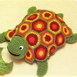 May include: A crocheted turtle toy with a green shell and a colorful, geometric pattern on its back. The turtle has a smiling face with black eyes and a black mouth.