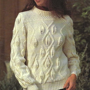 Puede incluir: Un suéter de punto blanco con un patrón de punto de cable y agujeros en forma de hoja. El suéter tiene cuello redondo y mangas largas.