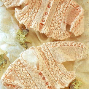 以下が含まれることがあります： ボタン留めとテクスチャードデザインのピーチカラーのニットベビーカーディガン2枚。カーディガンはフリルのあるヘムがあり、薄い茶色の表面に置かれています。