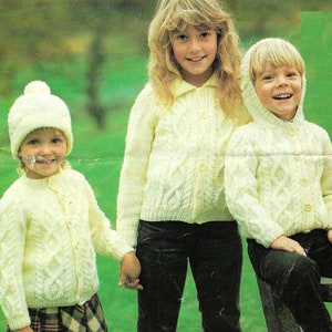 Pode incluir: Três crianças vestindo cardigãs de tricô branco com padrões de trança. A menina à esquerda está usando um chapéu de tricô branco com um pompom. A menina do meio está usando calças pretas. O menino à direita está usando um cardigã com capuz.