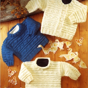 May include: Three knitted sweaters in cream and blue, with textured patterns. One cream cardigan has button closures. Decorative paper cutouts of geese and chickens are scattered around the sweaters on a wooden surface. The sweaters are likely for children.