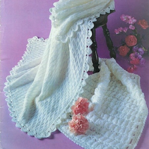 Könnte beinhalten: Cremefarbene, gestrickte Babydecke mit gewelltem Rand und Rautenmuster. Die Decke ist über einen dunklen Holzstuhl drapiert, und eine zweite Decke ist darunter gefaltet. Zwei rosa Nelken sind auf der Decke platziert.