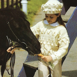 Può includere: Una giovane persona indossa un maglione bianco lavorato a maglia con un motivo testurizzato e un cappello bianco lavorato a maglia e nutre un cavallo nero con una carota.