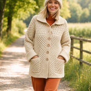 May include: A beige knit cardigan featuring a textured diamond pattern, a wide collar, long sleeves with folded cuffs, and two front pockets. The garment is fastened with a vertical row of five round buttons. A matching beige knit beanie is also worn.