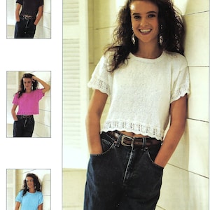 May include: A woman is wearing a white crocheted top with a scalloped hem and blue jeans. She is standing in front of a white wall with a window behind her.